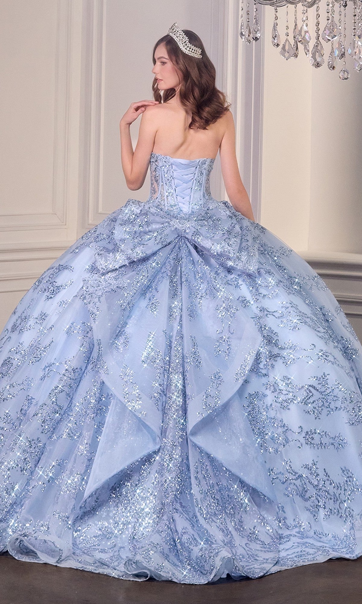 A woman wearing the Quinceanera Dress 15732 By Ladivine—a strapless ball gown with a corset bodice and glitter details—stands in an elegant room with white walls, a chandelier, and a sparkling tiara.
